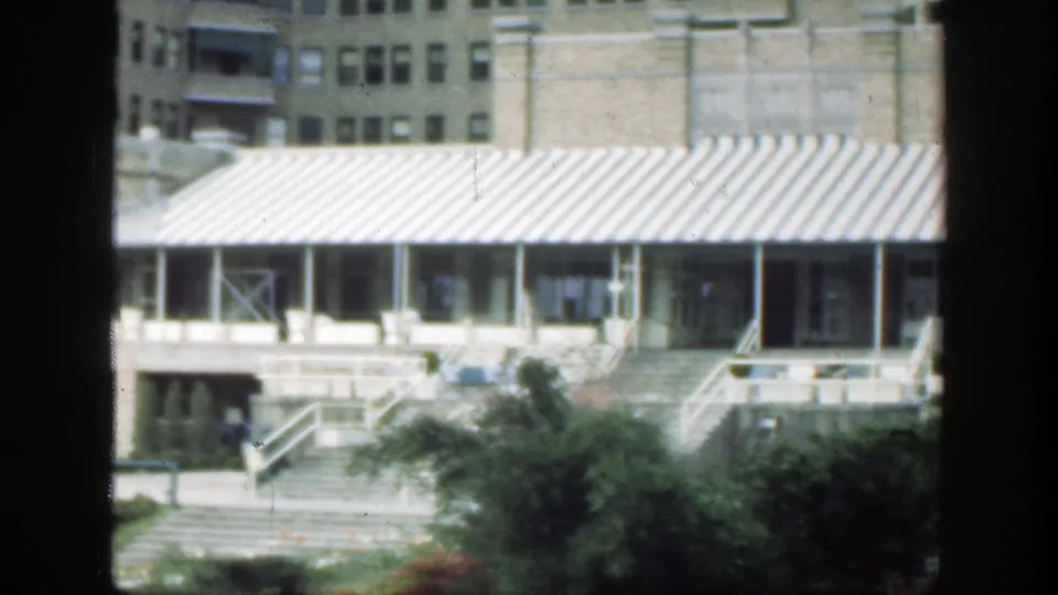 1949 Cincinnati home-movie frame showing the awning-covered entrance, steps, landscaped shrubs, upper masonry wall, and small distant figures near the building.