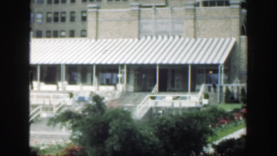 1949 Cincinnati 8mm frame showing a brick facade with a striped awning, front steps, garden beds, and shrubs viewed through a window frame.