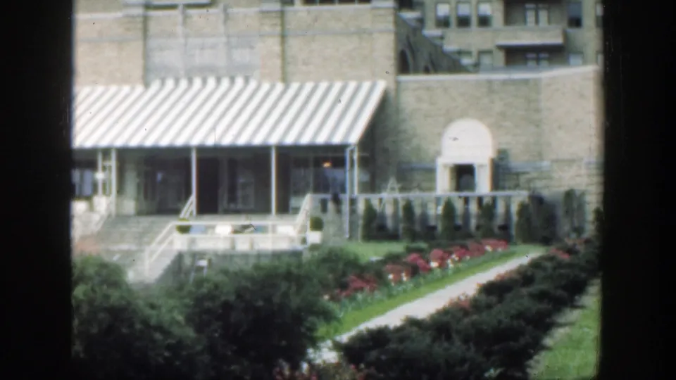 Soft 1949 Cincinnati 8mm view of an awning-covered building entrance with nearby arched doorway, steps, hedges, red flowers, and lawn.