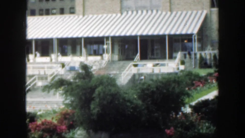 1949 Cincinnati 8mm frame showing a striped awning over a brick facade, entry steps, shrubs, lawn, and flowers behind a dark window edge.