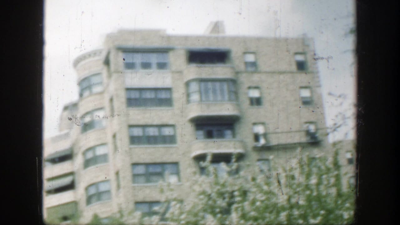 Pale multi-story building seen through dark window framing with curved corner bays and trees below.
