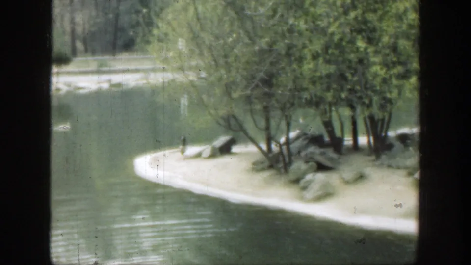 1949 Ohio home-movie frame showing a curved sandy island edge, trees, rocks, and calm pond water.