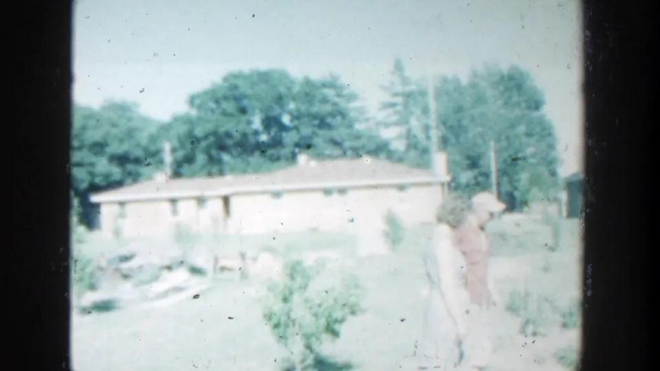 In Wisconsin home-movie footage from 1964, two older adults stand in a yard beside a low house and garden growth.