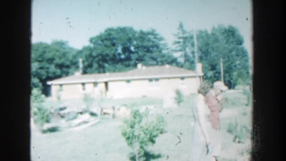 Two older adults stand near a garden area outside a modest Wisconsin house in soft, faded 1964 8mm footage.