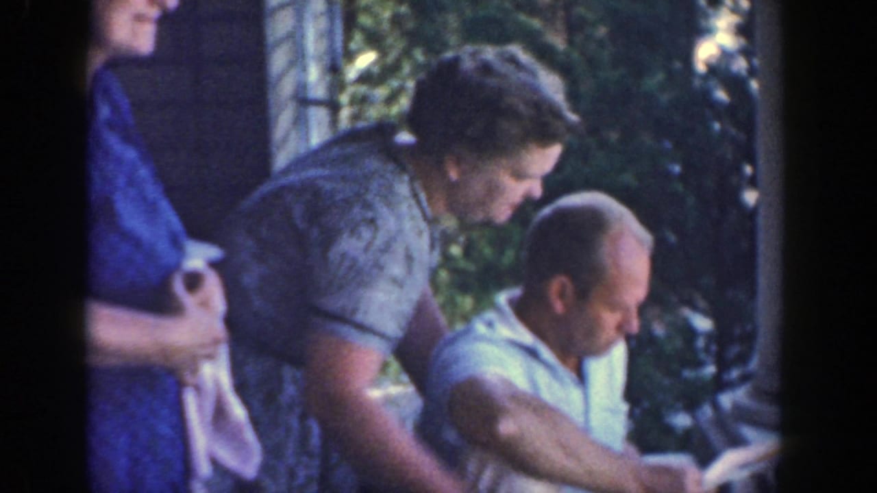 Adults gather on a porch as a seated man looks down and two women stand nearby.