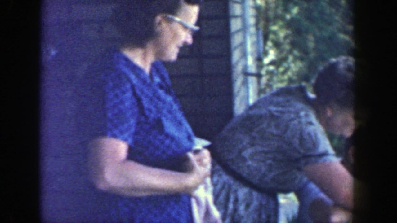 A woman in a blue patterned dress and glasses stands by a brick porch as another woman leans forward beside her.