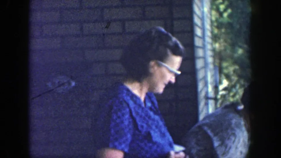 The home-movie frame centers on a woman in a blue patterned dress and glasses standing on a Wisconsin porch.