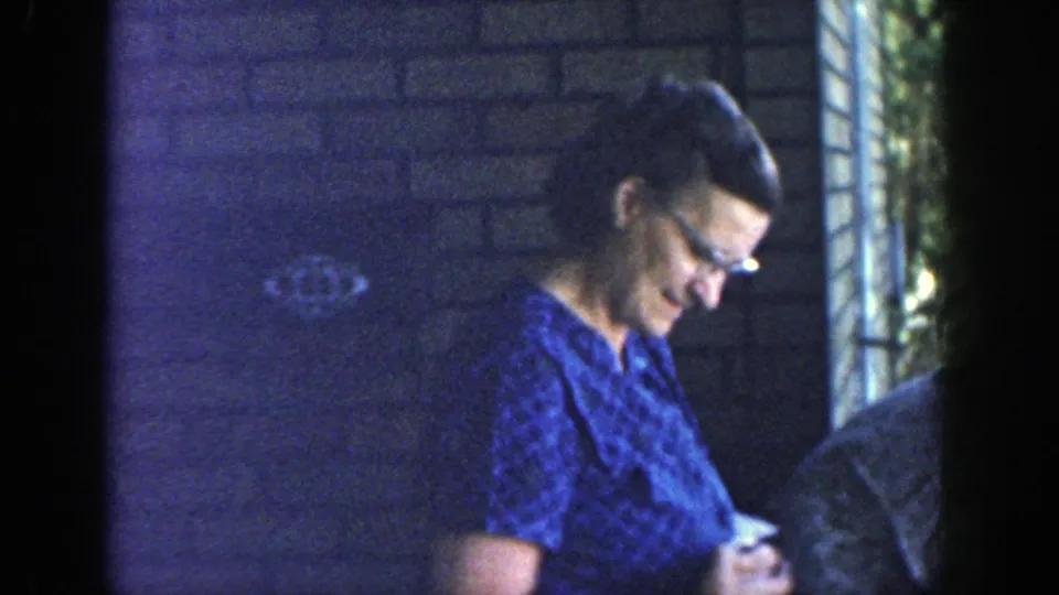 A woman in a blue patterned dress smiles on the porch, with brickwork, greenery, and another adult partly visible nearby.