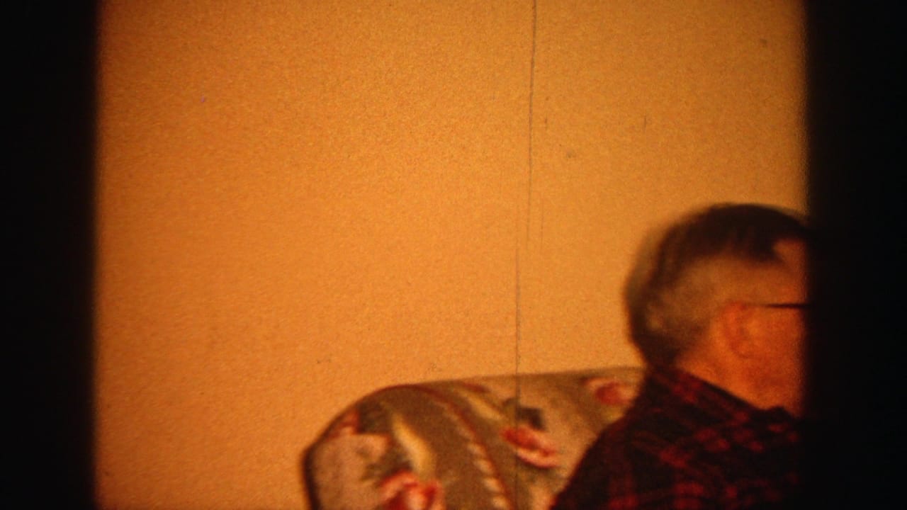 Older man in glasses seated on a floral sofa in a warm living room.