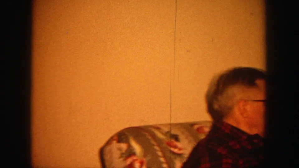 In a 1964 Wisconsin home-movie frame, an older man in glasses sits on a floral sofa in a warmly lit living room.