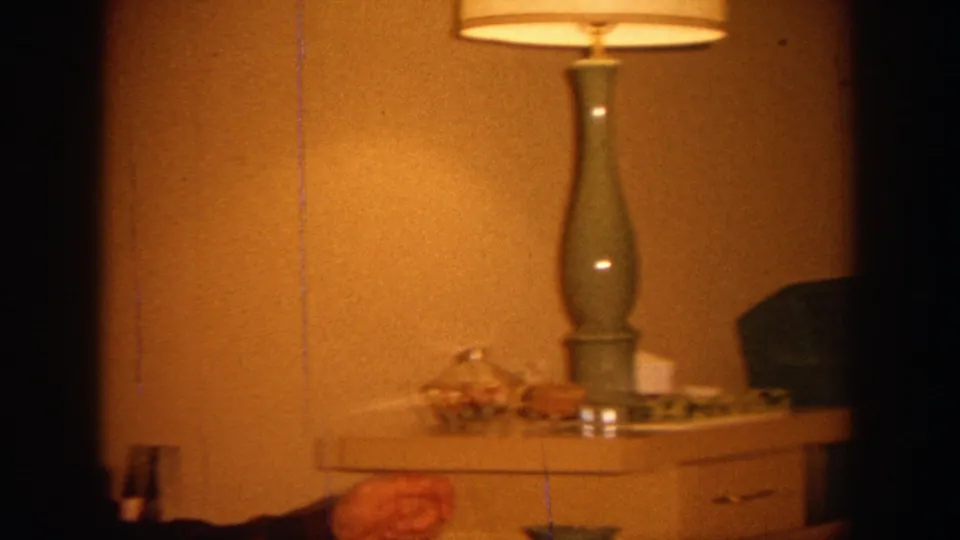 A warm 8mm frame shows a table lamp, side table, green chair, and a partially visible seated person in the living room.