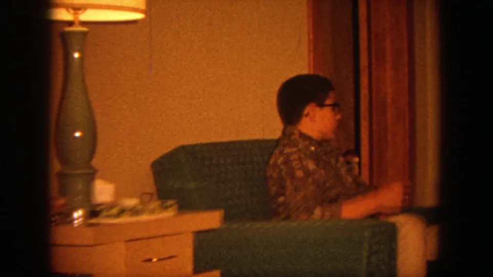 A young person with glasses sits in a green chair near curtains and a side table in the 1964 Wisconsin living room.