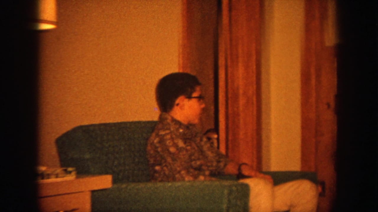 Boy with glasses sits in a green armchair beside a woman at a kitchen table.