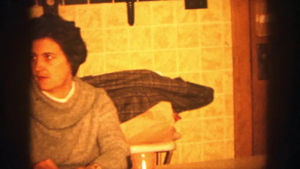 1964 Wisconsin home-movie frame of a seated woman in a gray cowl-neck sweater at a warm kitchen table.