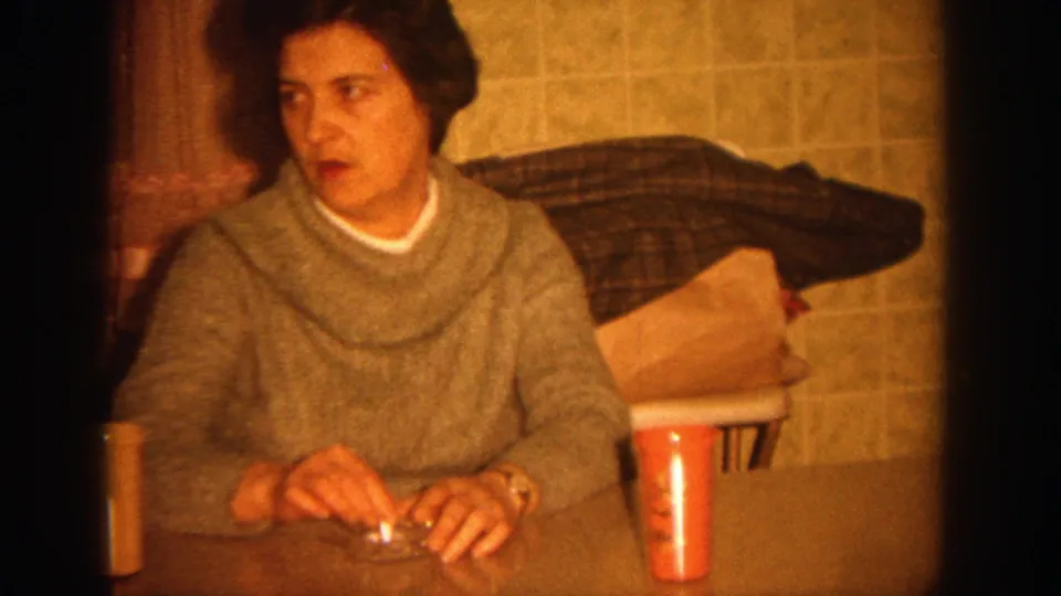 1964 Wisconsin home-movie frame showing a woman seated at a kitchen table in warm 8mm light.