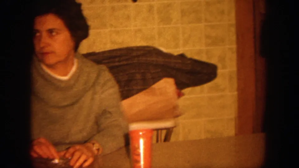 1964 Wisconsin home-movie frame of a woman seated in a tiled kitchen, looking slightly to the side.