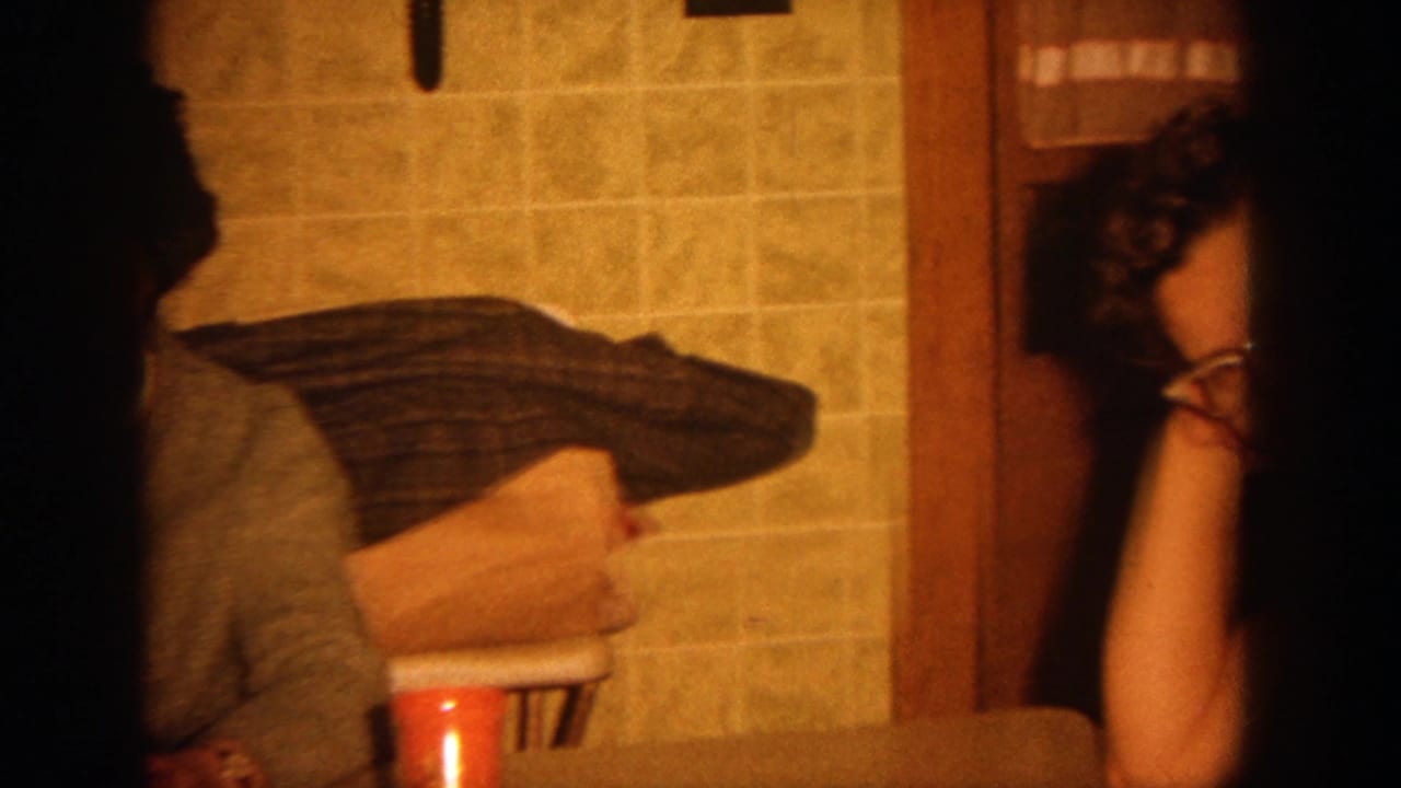 1964 Wisconsin home-movie frame showing a partially visible adult woman in a warm kitchen with yellow tile, folded clothing, and a red cup.