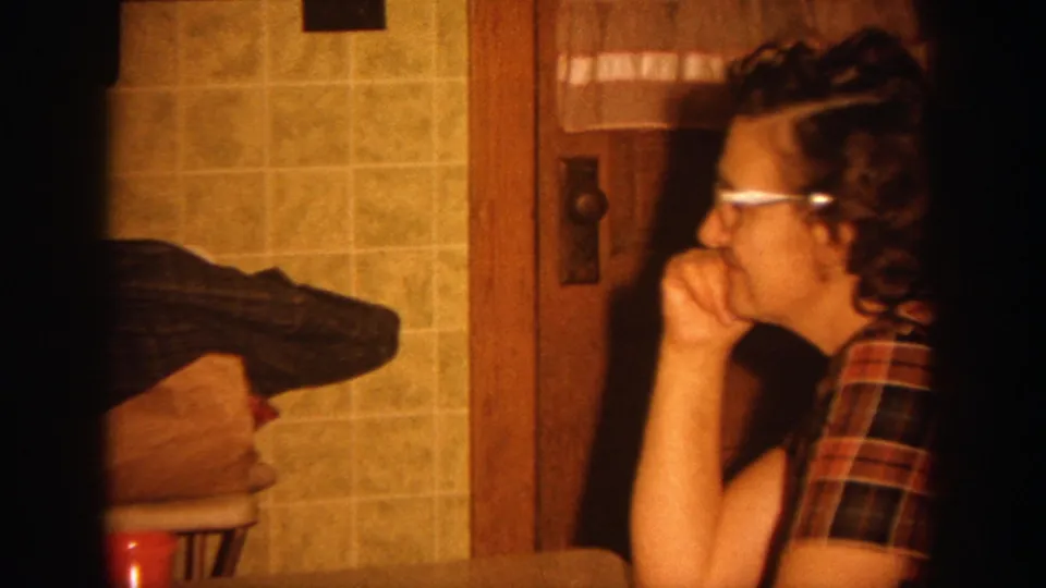 1964 Wisconsin home-movie frame of an adult woman in glasses and a plaid shirt seated in a warmly lit kitchen.