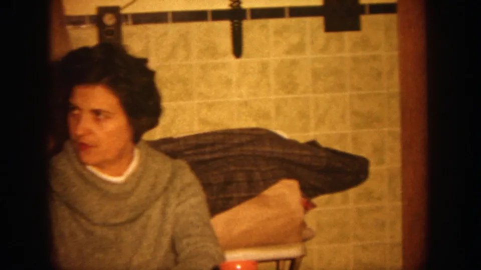1964 Wisconsin home-movie frame showing an adult woman in a gray cowl-neck sweater seated at a kitchen table.