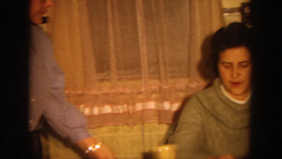1964 Wisconsin home-movie frame of a woman seated near kitchen curtains, with another adult partly visible at the table.