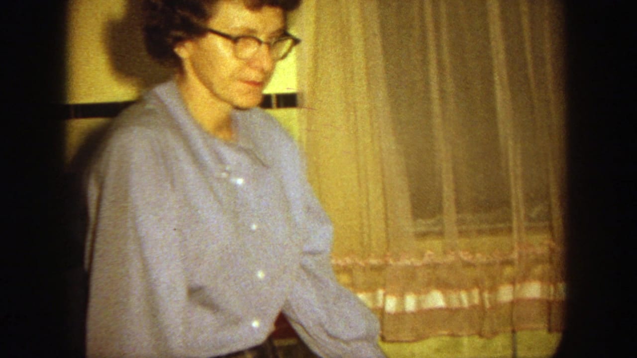 Woman in glasses and a blue shirt stands indoors near curtains and tiled wall trim.