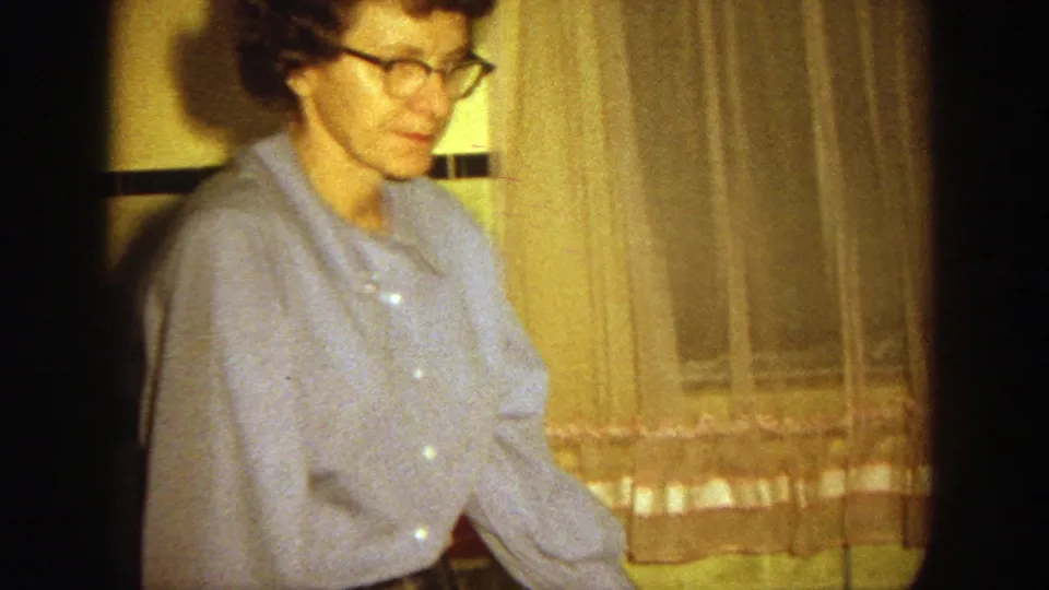 In 1964 Wisconsin home-movie footage, a woman in glasses and a blue shirt stands in a warmly lit interior near curtains.