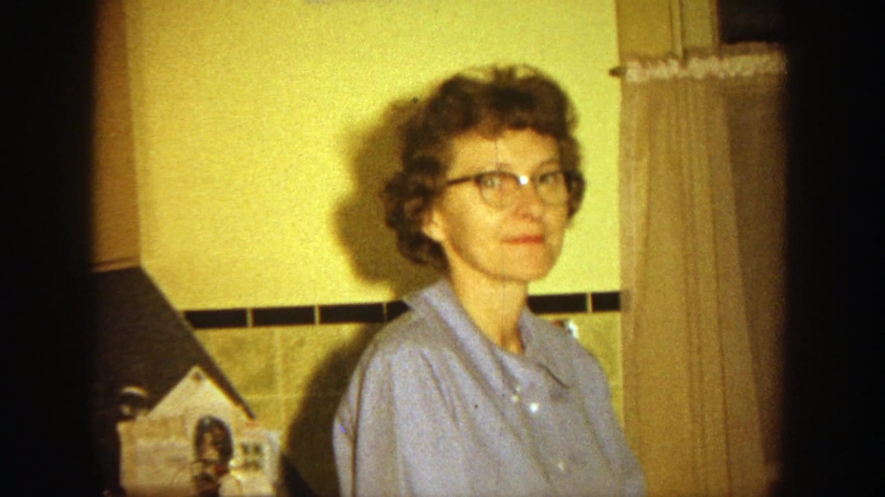 Woman in glasses and a blue shirt faces the camera beside curtains and wall decor.