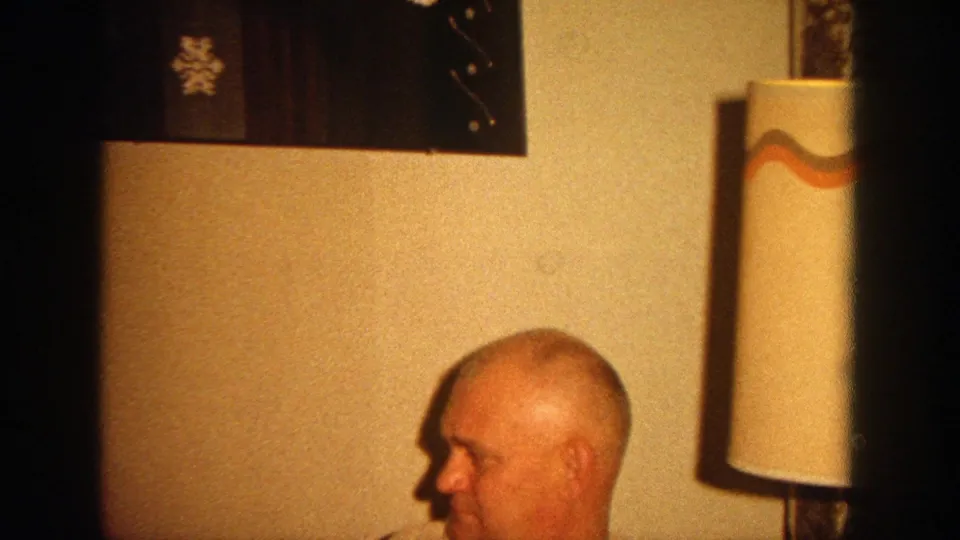 A seated bald man appears in the lower frame beneath a lamp and a Merry Christmas wall decoration in a private interior.