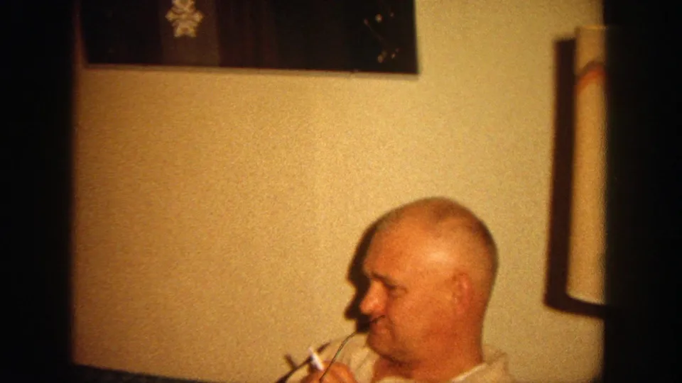 The seated man smokes a cigarette beside a lamp and wall decoration in the warmly lit 1964 Wisconsin home-movie scene.