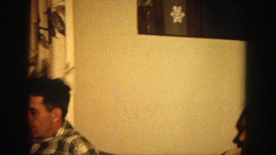The camera shifts across the living room, showing the plaid-shirted man partly cropped beside wall decor and a curtained doorway.