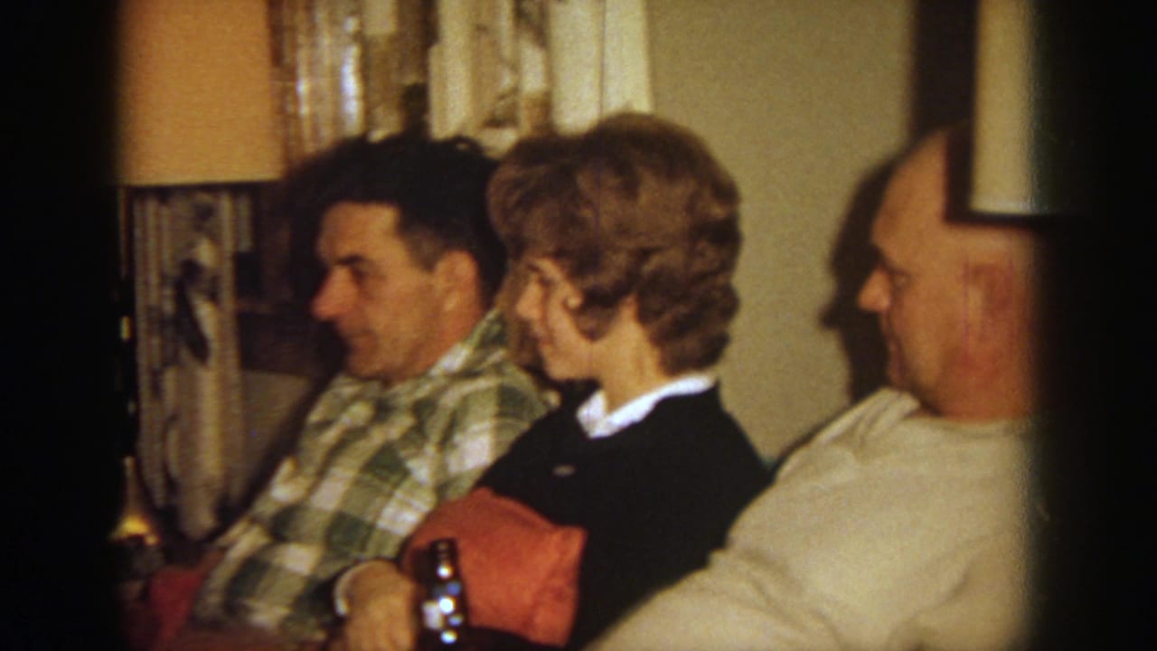 Three adults seated indoors during a family gathering, with a plaid shirt, dark sweater, and red cushion visible.