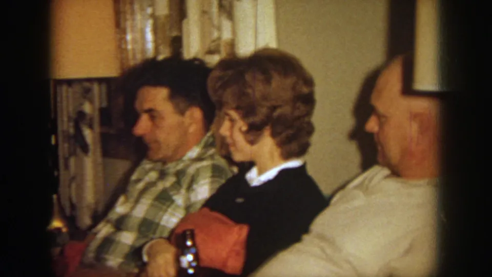 1964 Wisconsin home-movie frame showing three adults seated closely in a living room during an informal family gathering.