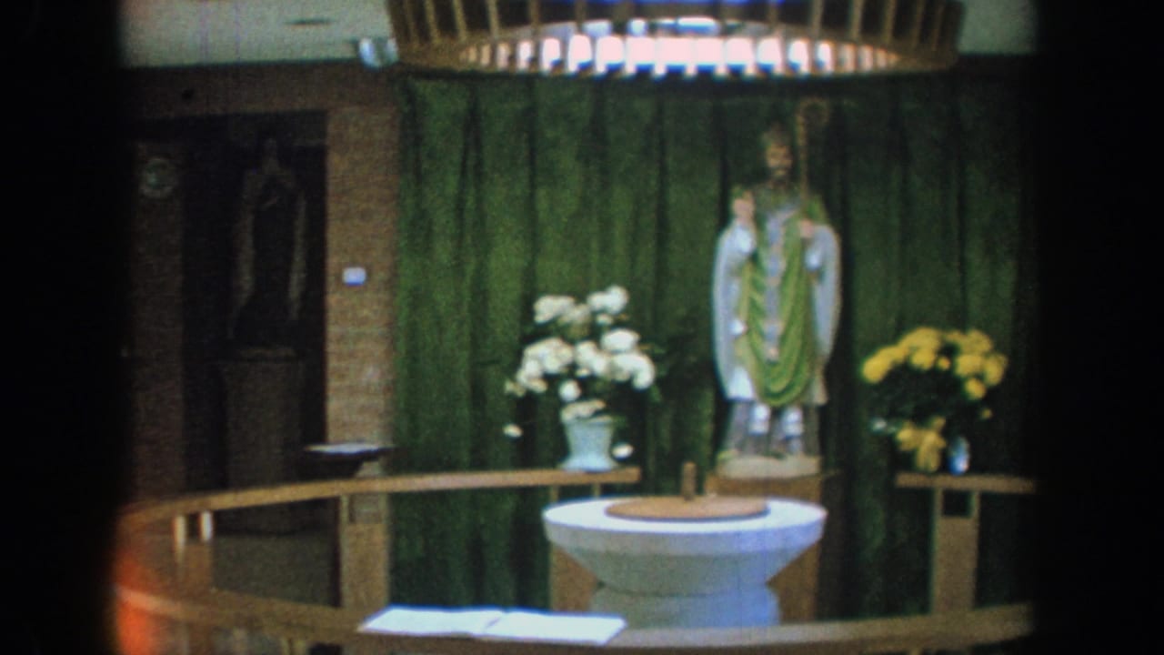Church interior with baptismal font, saint statue, green drapery, flowers, railing, and brick wall.