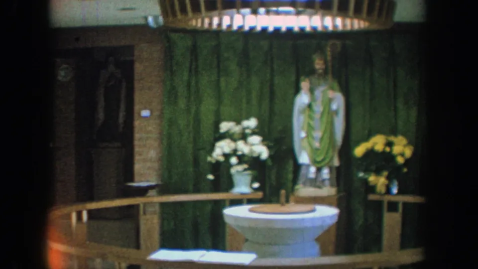 1964 Wisconsin church interior with a baptismal font before green drapery, floral arrangements, and a saint statue.
