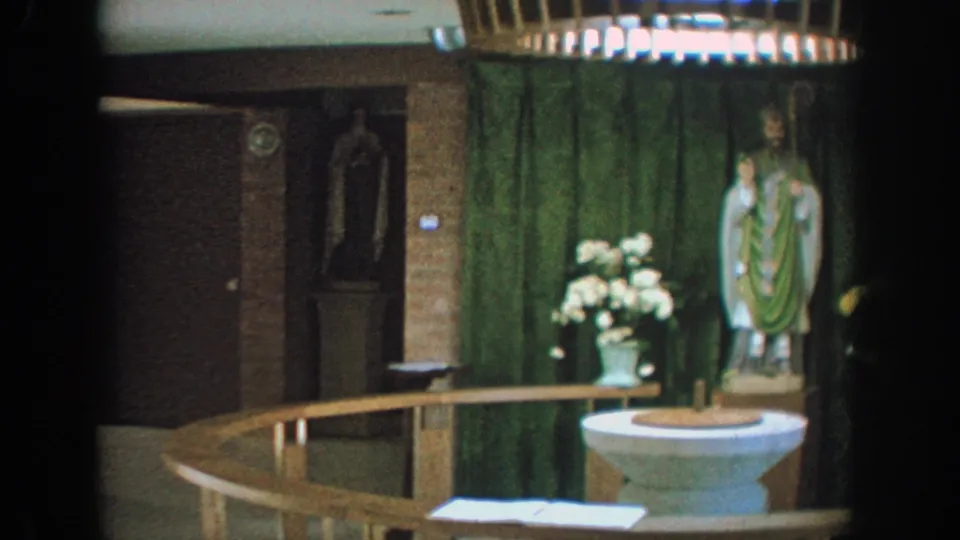 1964 Wisconsin home-movie frame of a church baptismal area with flowers, green drapery, saint statue, and railing.