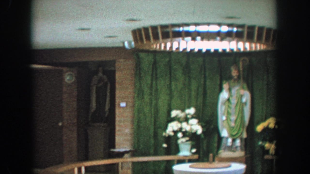 Interior religious display with saint statue, green curtain, flowers, and white font.