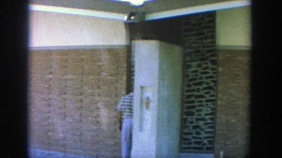 1964 Wisconsin 8mm frame showing a man partly turned away near a white door panel with a cross, framed by tan brick and dark stonework.