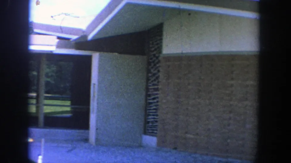 1964 Wisconsin 8mm frame of a covered exterior walkway beside tan brick and dark stone walls, with an open grassy area visible in the distance.