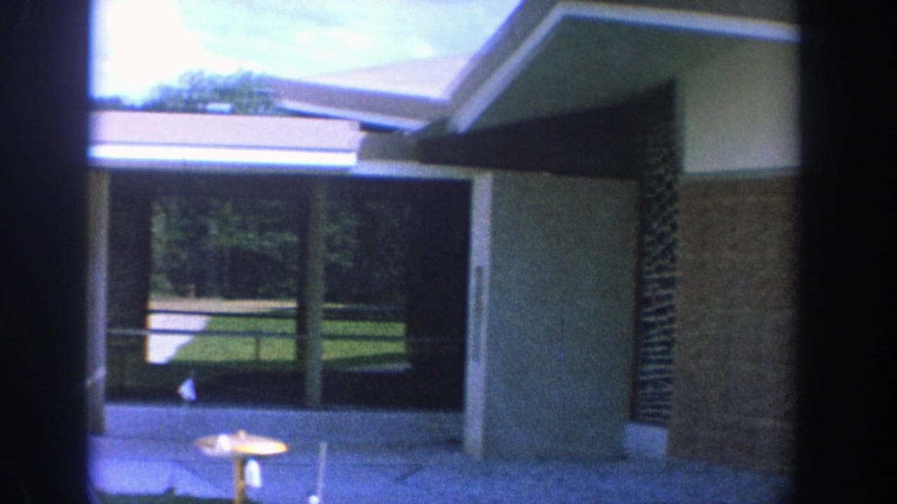 Modernist building exterior with large windows, walkway, lawn, and a small round object near the entrance.
