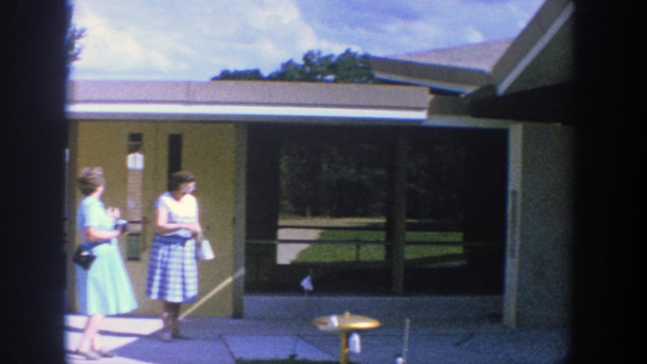 Two adult women stand near a building entrance, one in a light blue dress and one in a checkered dress.
