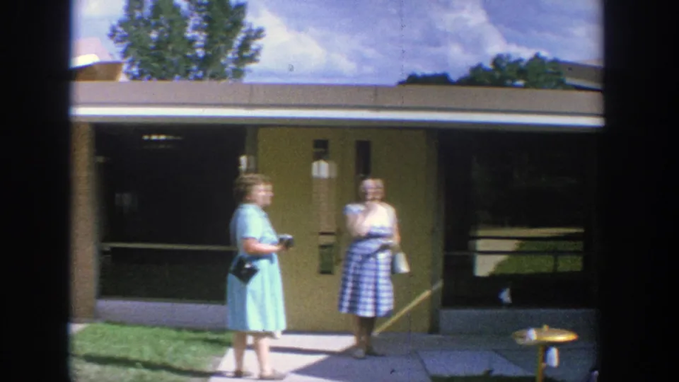 1964 Wisconsin frame showing two adult women talking on a walkway outside a flat-roof building with dark windows.