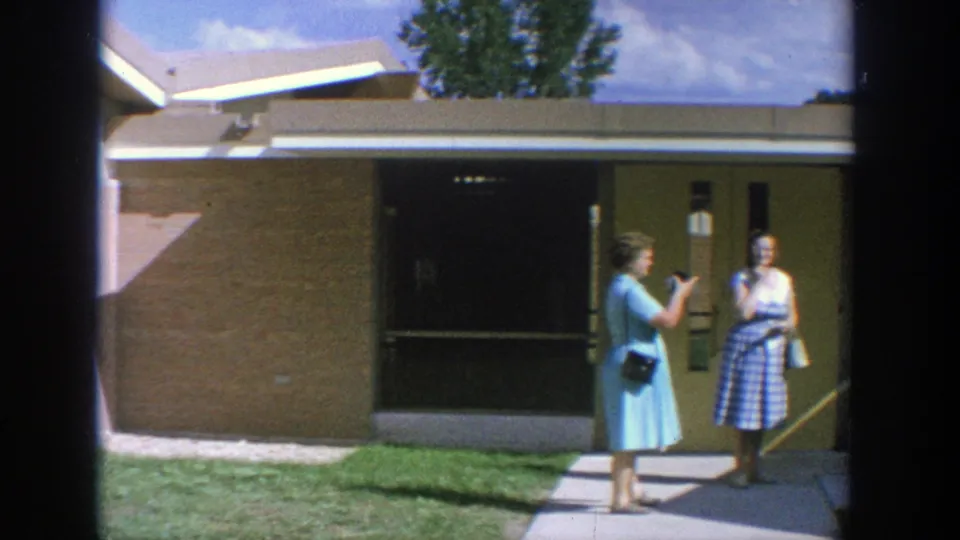 1964 Wisconsin 8mm frame of two adult women with handbags conversing outside a modernist building facade.