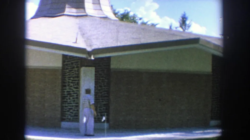 1964 Wisconsin home-movie frame showing an adult near the brick entrance column of a mid-century church or tabernacle.