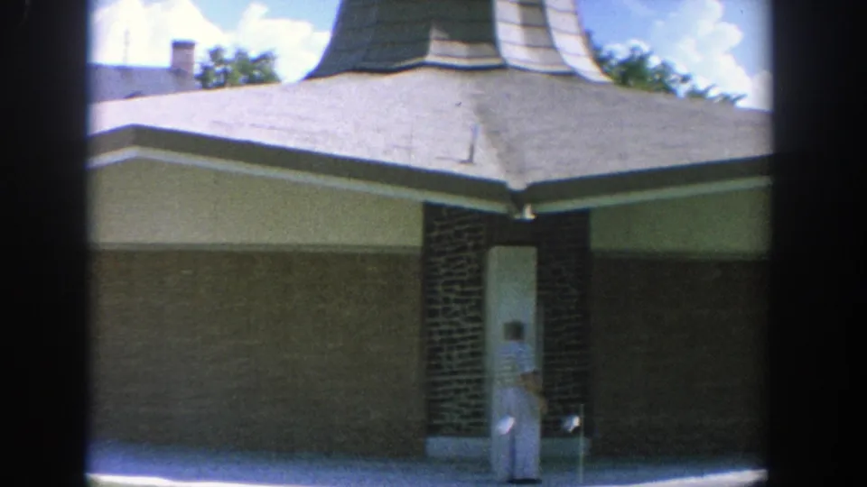 1964 Wisconsin frame of a man walking near the entrance of a mid-century church or tabernacle exterior.