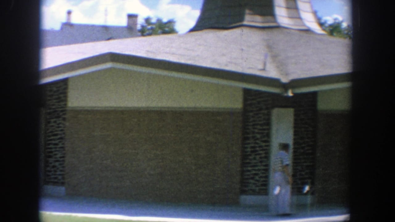 1964 Wisconsin 8mm frame showing two women in blue and white dresses near a mid-century building exterior.