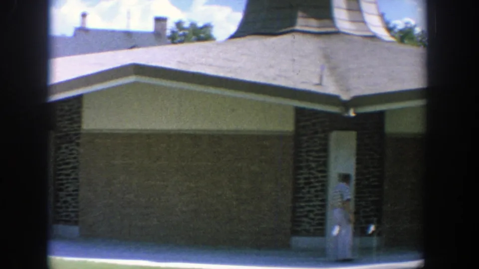 1964 Wisconsin 8mm frame showing two women in blue and white dresses near a mid-century building exterior.