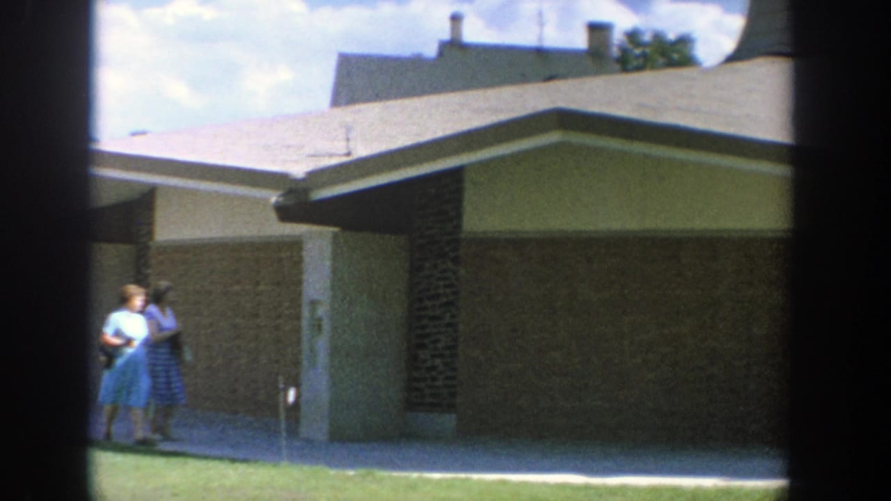 Two women walk beside a low building with brick walls and a grassy lawn.