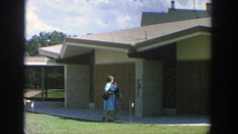 1964 Wisconsin 8mm frame showing a woman in a blue dress near a sidewalk, lawn, and mid-century building exterior.