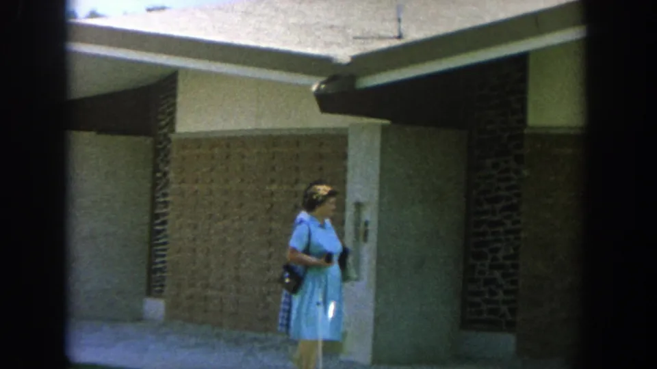 1964 Wisconsin home-movie frame of a woman in a blue dress near a doorway, brick wall, and stucco exterior.