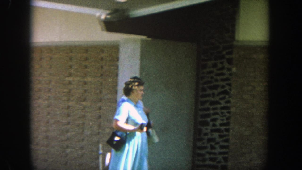 1964 Wisconsin 8mm frame showing an adult woman in a light blue dress walking past a tan brick exterior with dark stone trim.